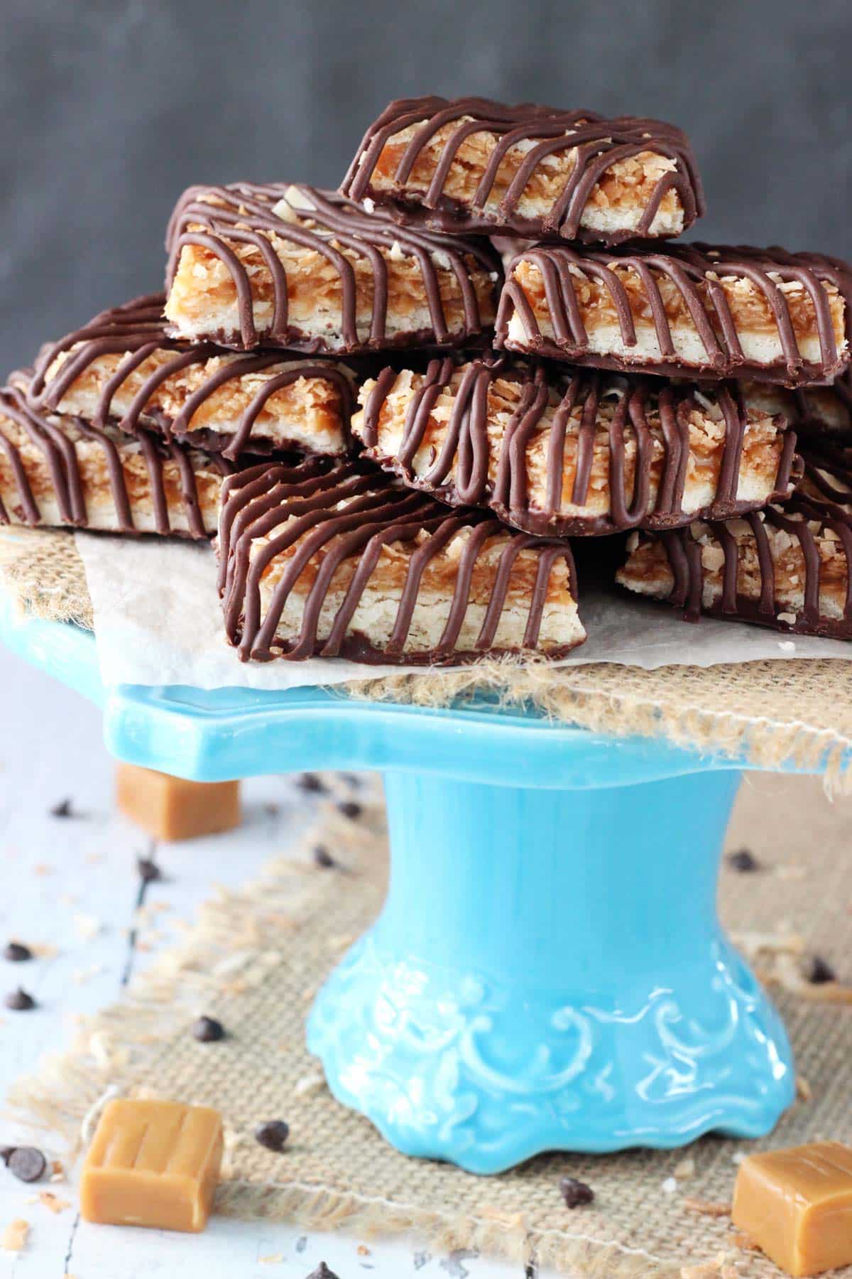 Buttery shortbread cookies are topped with gooey caramel, nutty toasted coconut and rich chocolate in these heavenly Samoa Cookie Bars! A blue cake stand topped with cut Samoa Cookie Bars.