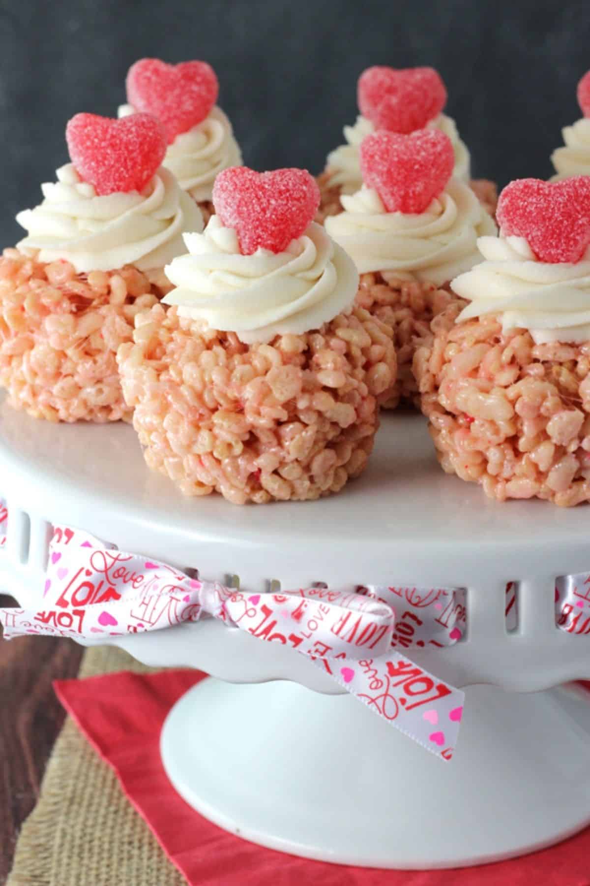 Several Valentine's Day rice krispie treats on top of a white cake stand tied with a valentine's ribbon.