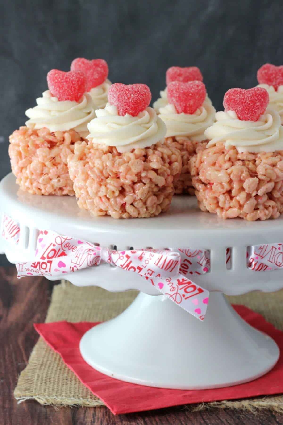 Several Valentine's Day rice krispie treats on top of a white cake stand tied with a Valentine's ribbon.