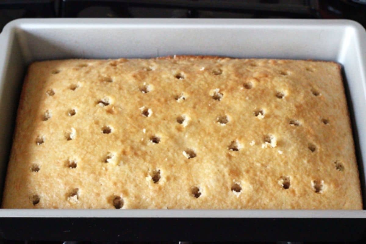 A yellow cake in a metal baking pan with holes poked into it.