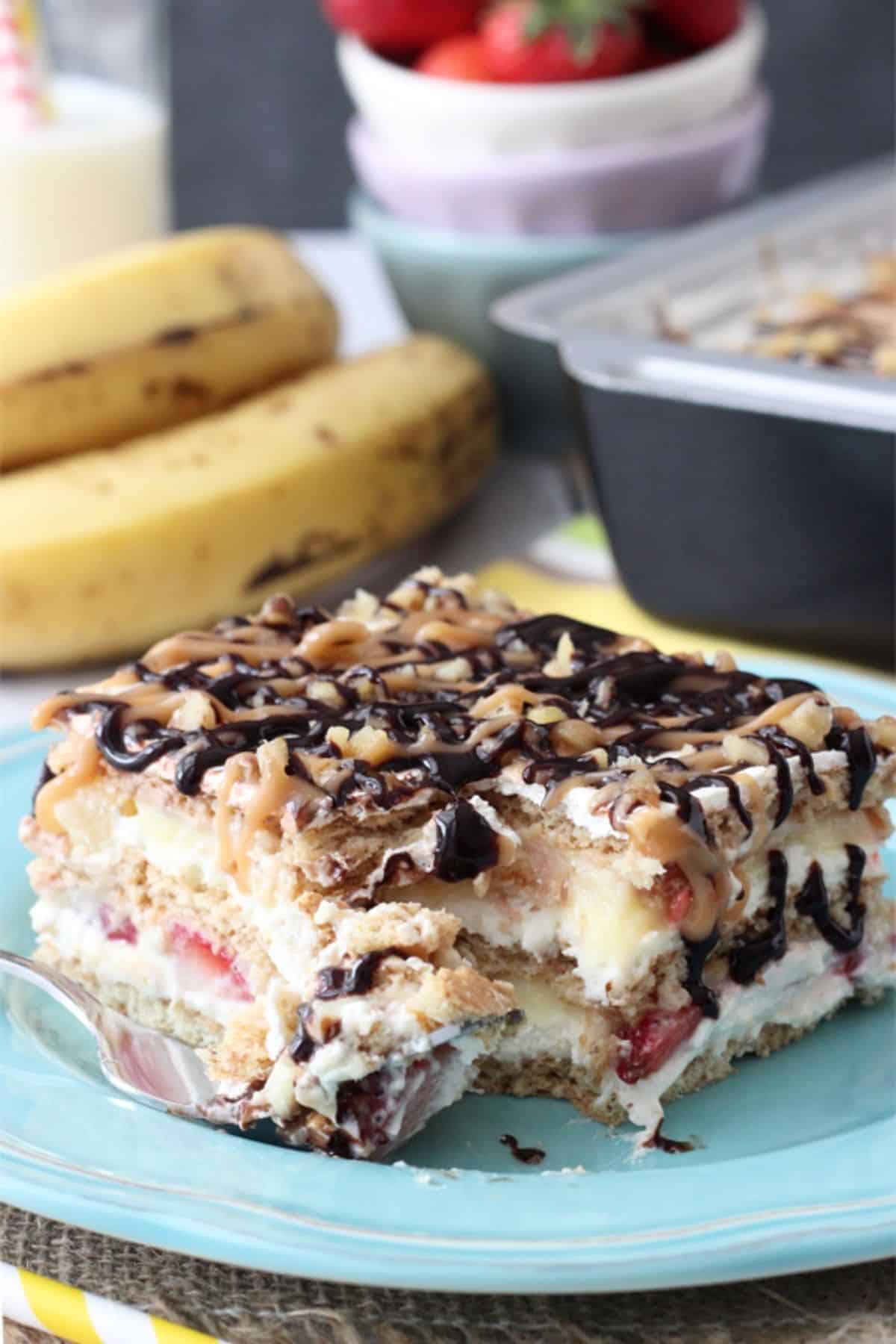 A slice of banana split cake on a blue plate with a bite removed on a fork.