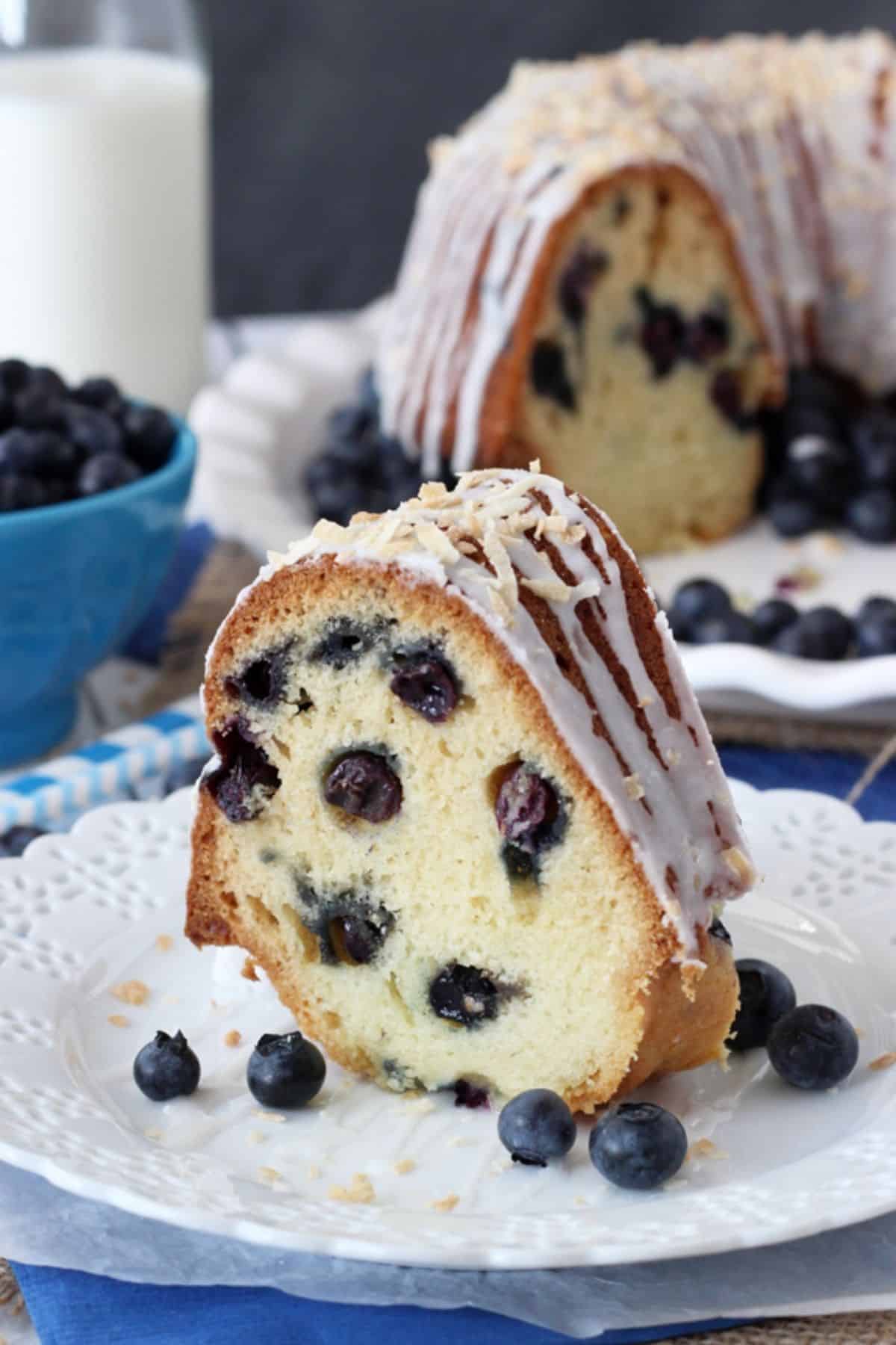 A slice of blueberry coconut bundt cake on a white plate with the rest of the cake in the background.