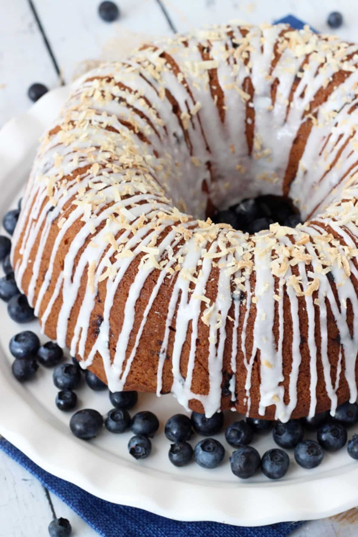 A glazed blueberry coconut bundt cake on a white cake plate surrounded by blueberries.