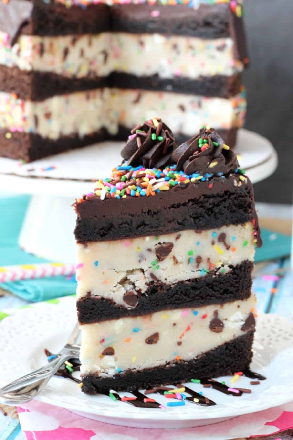 A tall slice of cookie dough brownie layer cake with the rest of the cake in the background.