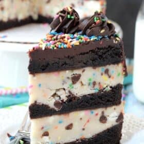 A tall slice of cookie dough brownie layer cake with the rest of the cake in the background.