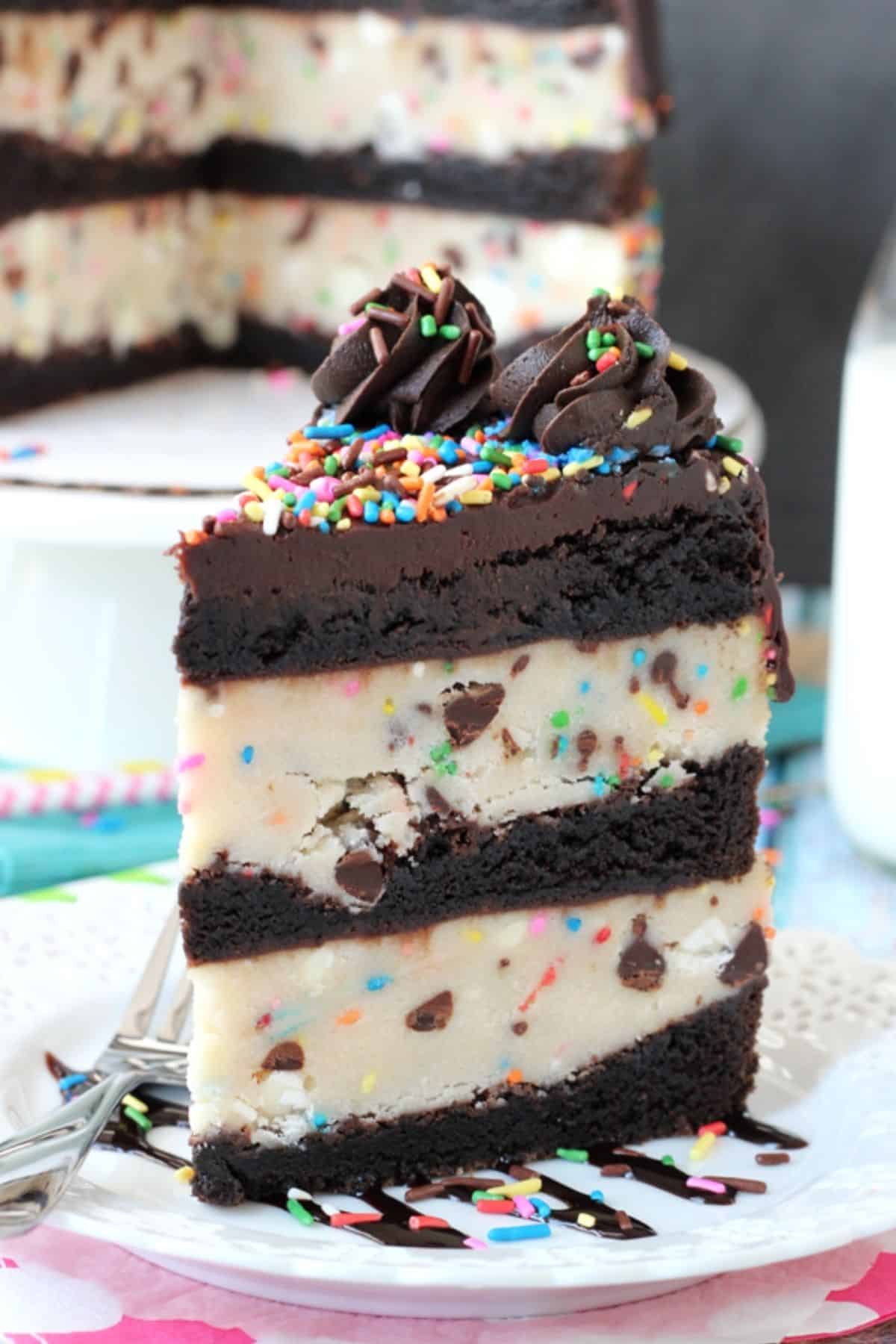 A tall slice of cookie dough brownie layer cake with the rest of the cake in the background.
