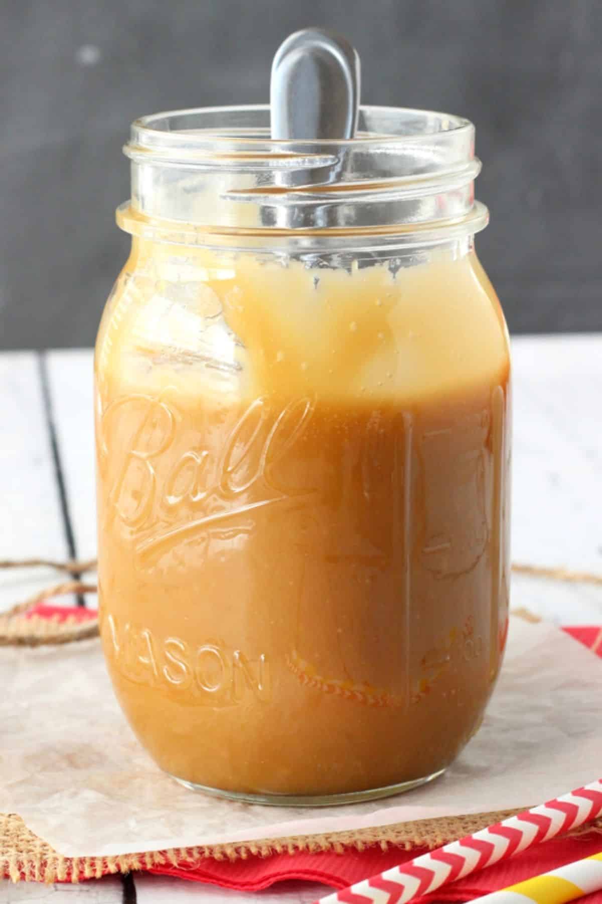 Close up of the side of a mason jar filled with caramel with a spoon inserted.