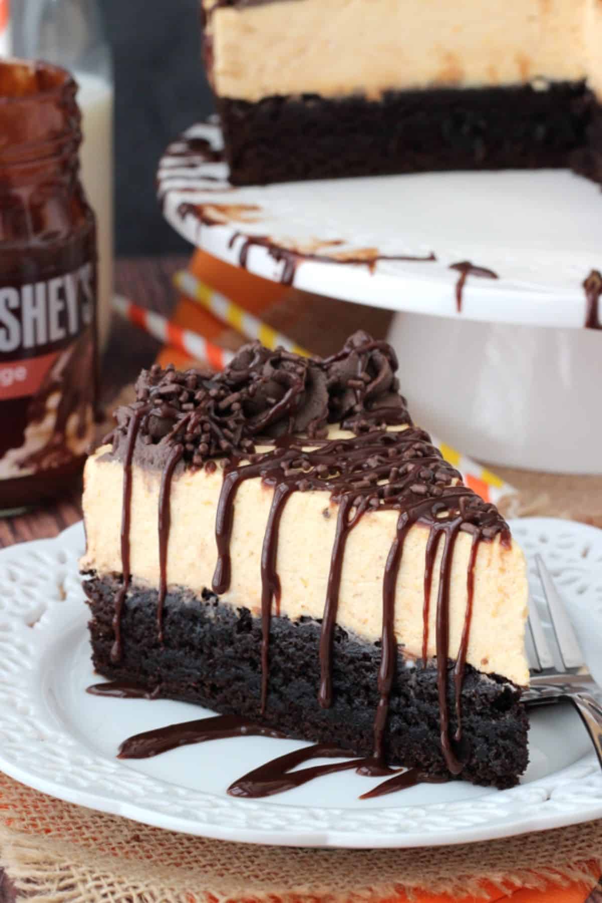 Close up of a slice of pumpkin chocolate brownie cheesecake on a small white plate with the rest of the cake in the background.