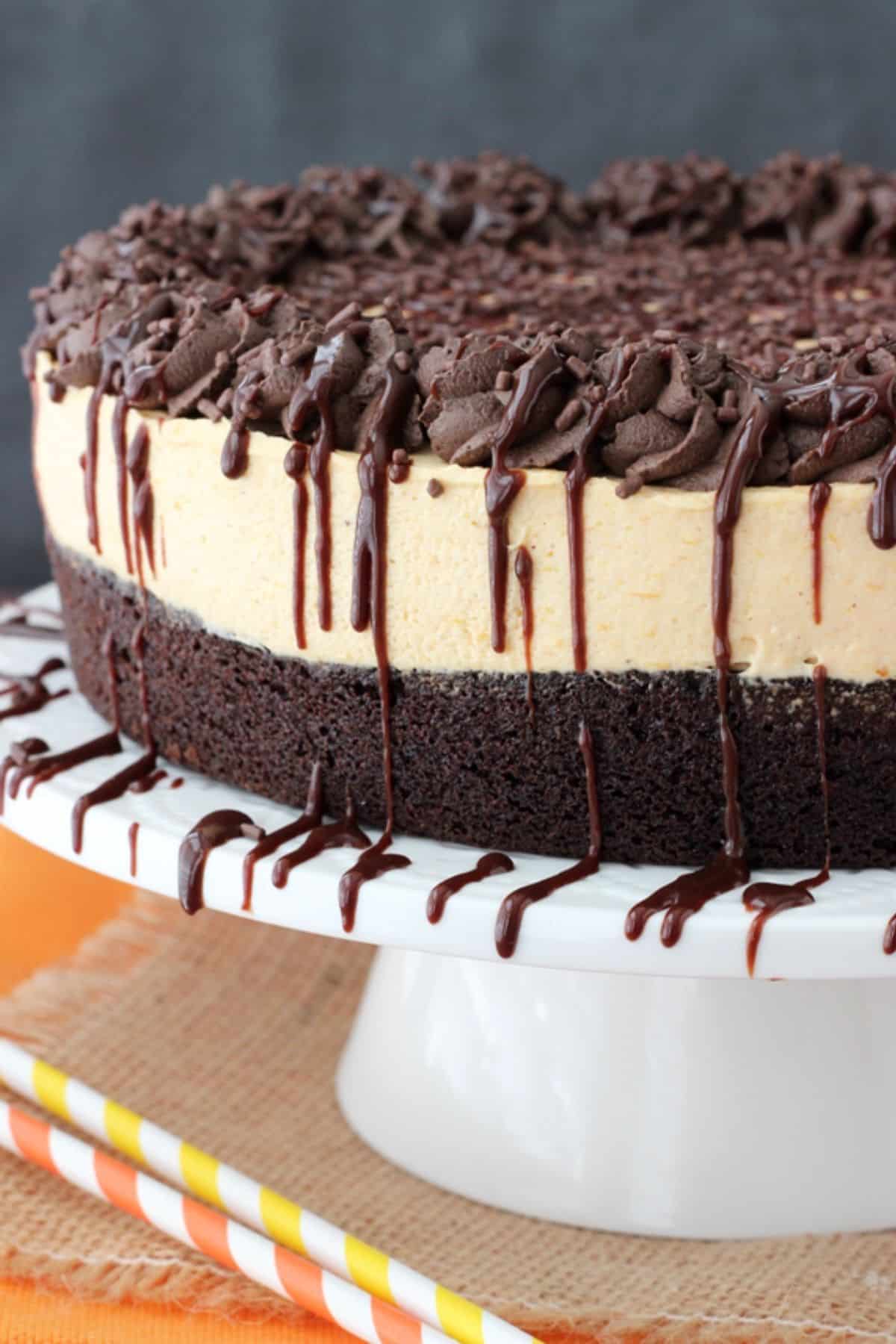 A pumpkin chocolate brownie cheesecake drizzled with chocolate on a white cake stand.