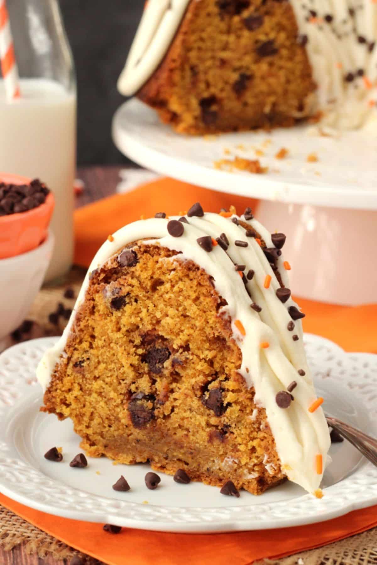 This pumpkin bundt cake with chocolate chips is soft, moist, and full of fall spices and gooey chocolate. It's perfect for Thanksgiving! Close up of a slice of pumpkin chocolate chip bundt cake on a small white plate with the rest of the cake in the background.