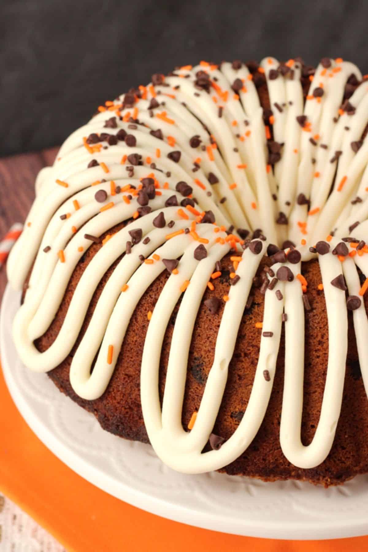 This pumpkin bundt cake with chocolate chips is soft, moist, and full of fall spices and gooey chocolate. It's perfect for Thanksgiving! Close up of a pumpkin chocolate chip bundt cake decorated with frosting and sprinkles on a white platter.