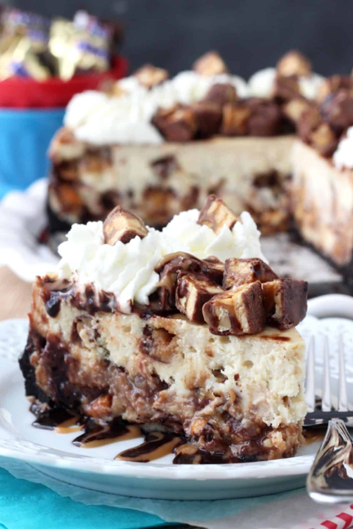 A slice of Snickers cheesecake on a small white plate with the rest of the cheesecake in the background.