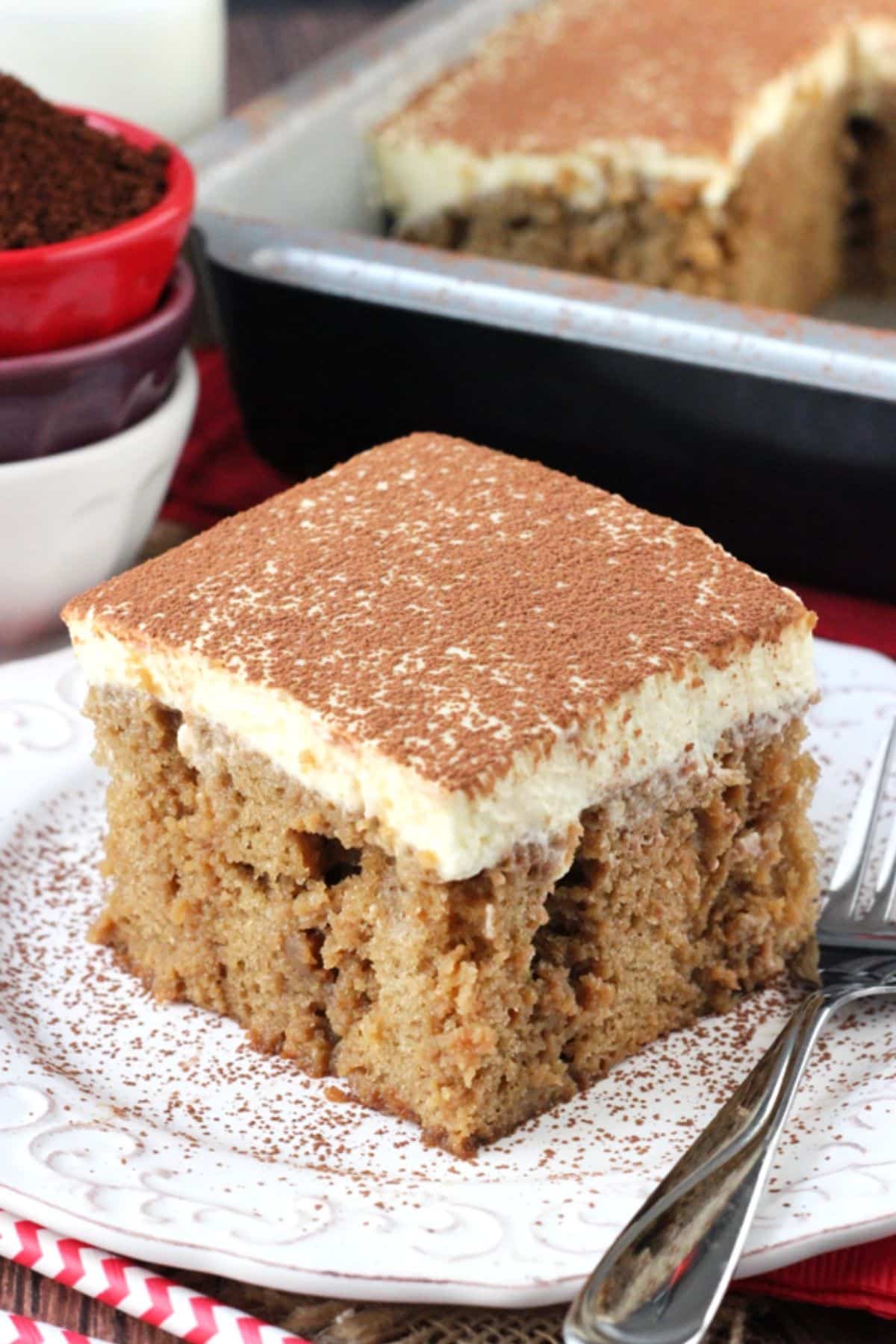 Close up of a slice of tiramisu poke cake on a small white plate with the rest of the cake in the background.