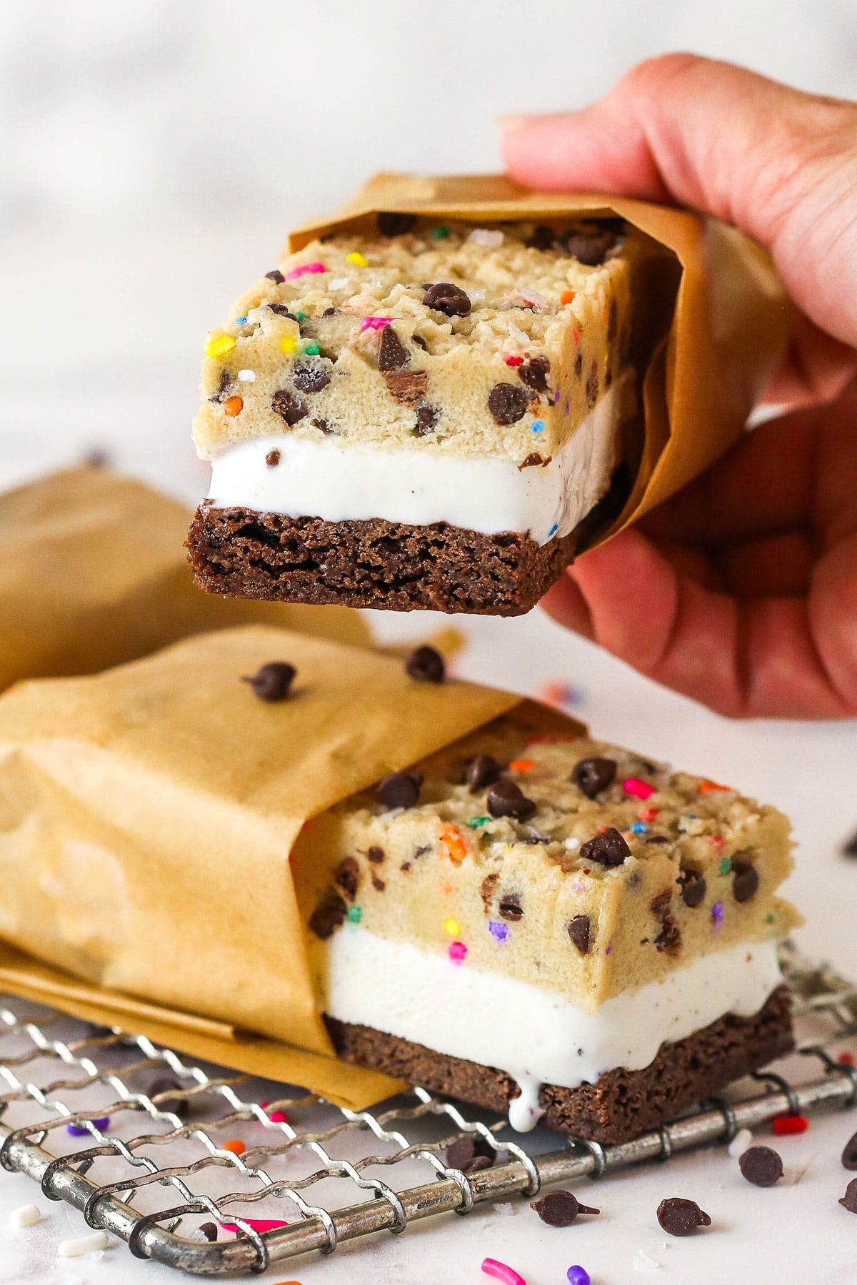 Brownie cookie dough ice cream sandwiches combine three favorites in one, with a few key ingredients to ensure they stay soft. A hand holding a brownie cookie dough ice cream sandwich over another one to show the three layers