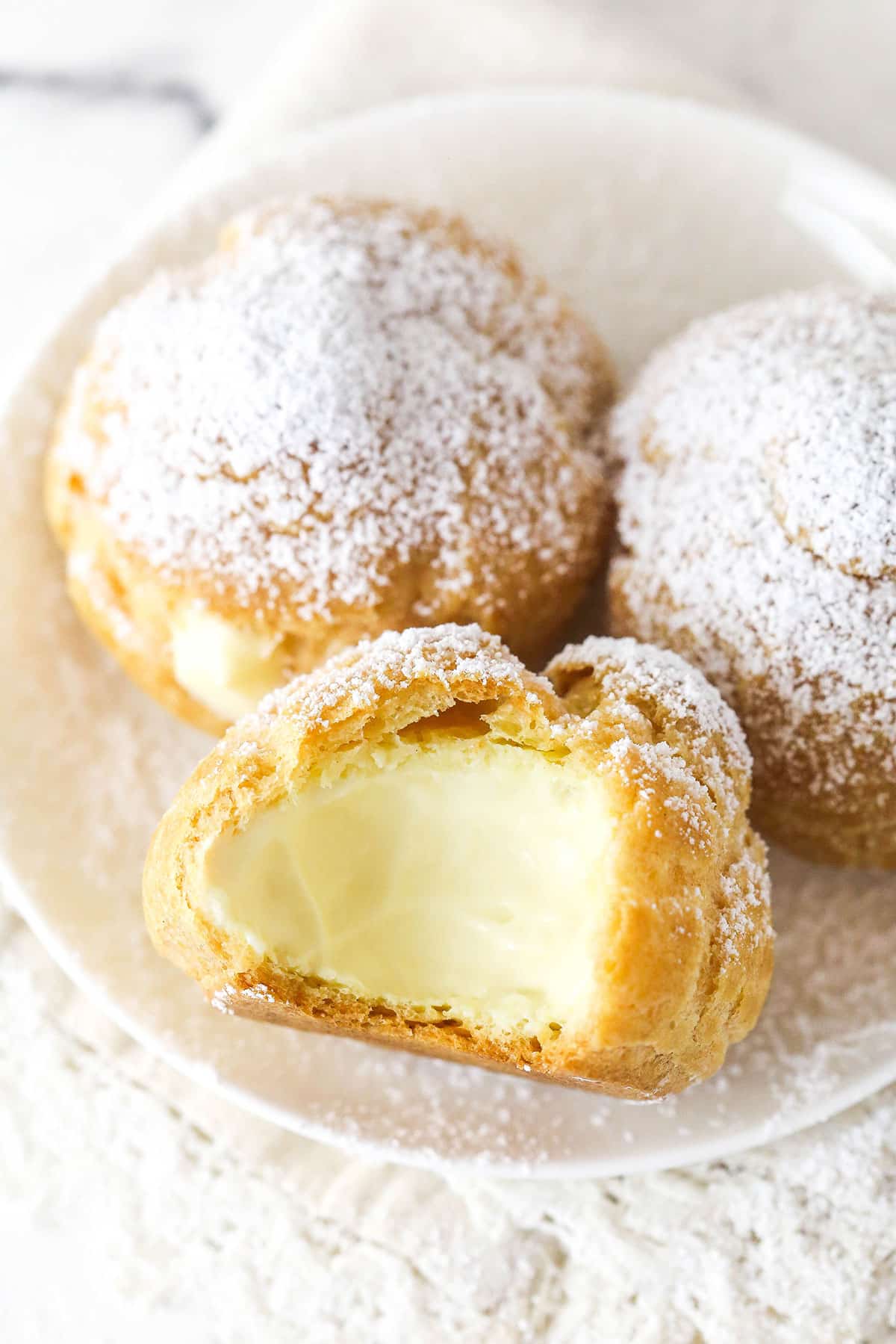 Master making cream puffs fit for a bakery and perfect for sharing with my in-depth tips and step-by-step photos. A plate of cream puffs, one with a bite missing to show the inside