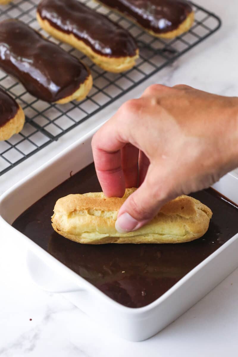 Mastering eclairs at home is easier than you think with my step-by-step photos and in-depth tips. Made with pastry cream & chocolate glaze. A hand dipping an eclair into melted chocolate