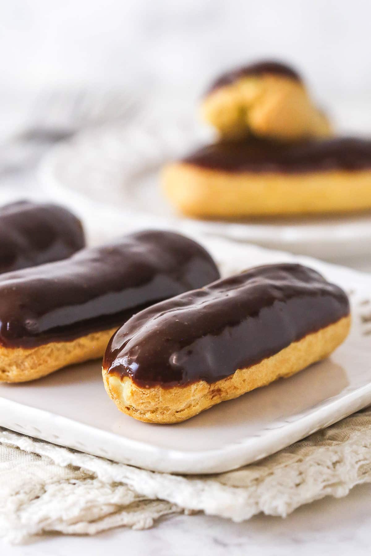 Mastering eclairs at home is easier than you think with my step-by-step photos and in-depth tips. Made with pastry cream & chocolate glaze. Chocolate eclairs on a white plate