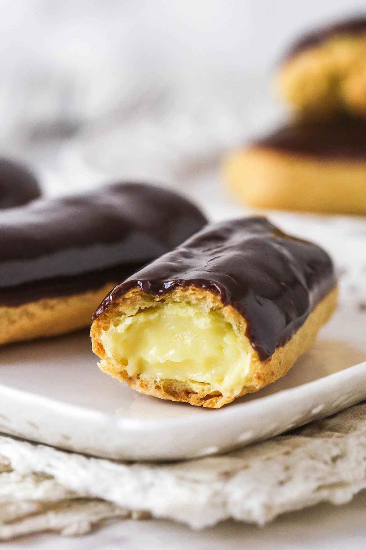 Mastering eclairs at home is easier than you think with my step-by-step photos and in-depth tips. Made with pastry cream & chocolate glaze. Homemade eclairs on a plate, one missing a bit to show the inside