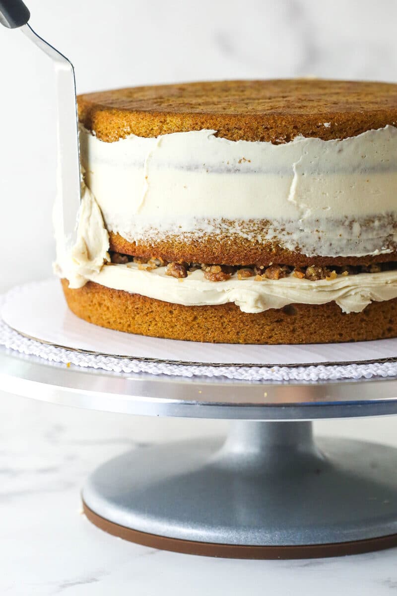 With layers of pumpkin cake, pecan praline crunch, and a caramel buttercream, this pecan praline pumpkin cake is impressive and delicious. A crumb layer being frosted on the outside of a pumpkin layer cake