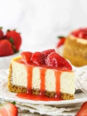 A slice of strawberry cheesecake on a plate with a fork near fresh strawberries and a full strawberry cheesecake.